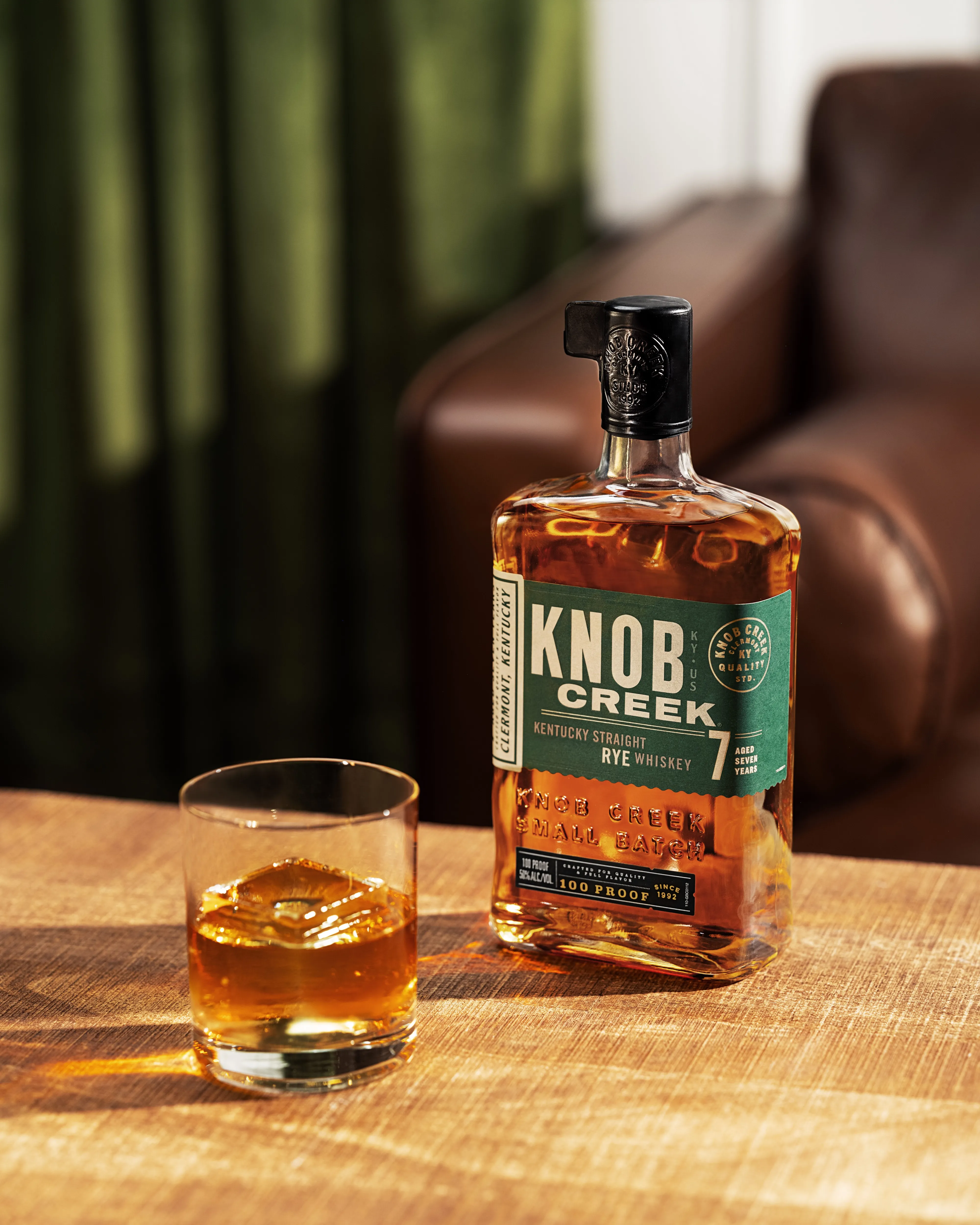 Bottle of knob creek whisky on table along with glass filled with same whisky.