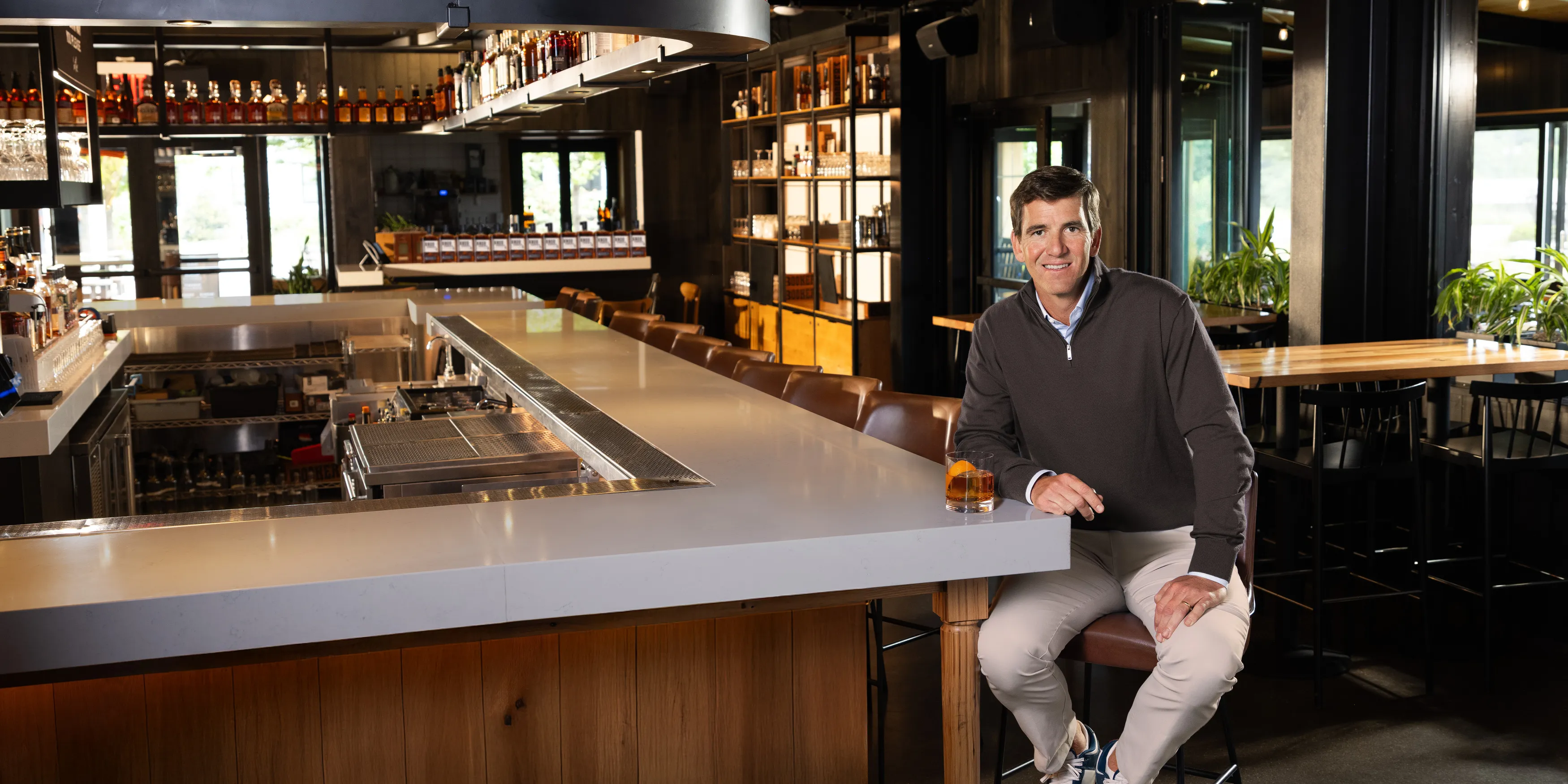 A man sits at a modern bar table with an orange cocktail in a stylish interior, featuring shelves lined with whiskey bottles.